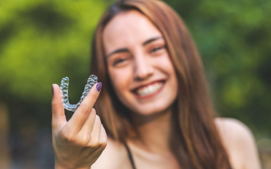 Image of a woman smiling holding a clear aligner - Invisalign braces - Orthodontics and Braces in Denver, Aurora, & Lakewood CO